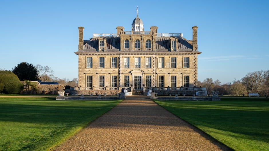 An elegant, historic stone mansion with a symmetrical facade featuring large rectangular sash windows, a central entrance with a decorative pediment, and a small dome topped with a flagpole on the roof. The building is surrounded by a well-maintained lawn with neatly trimmed grass, a gravel pathway leading directly to the front entrance, and low stone balustrades on either side of the stairs. In the background, there are a few trees with sparse foliage and a clear blue sky, indicating a bright, sunny day. The scene is characterized by a peaceful, manicured outdoor setting typical of estate grounds. This setting could relate to private or independent waste disposal activities, such as on-site clearance or external rubbish collection services, relevant to waste management and disposal discussions. The image emphasizes tidy, organized grounds consistent with professional waste handling practices.