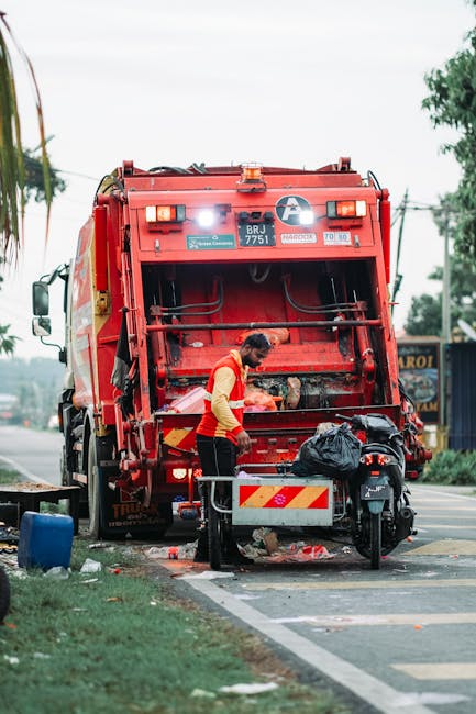 A worker in a yellow and red safety vest operating a large red rubbish collection truck on a roadside during daytime. The truck has a wide rear loading hatch open, revealing its internal mechanisms for waste collection. The worker is standing behind a small metal barrier attached to the truck, handling a black plastic bag filled with refuse, which is positioned near the truck’s rear platform. To the right of the worker, there is a motorcycle parked close to the truck, and scattered debris can be seen on the pavement underneath. The environment includes a grassy verge in the foreground, a few trees, and utility poles over a cloudy sky, indicating an urban or suburban street scene. The setting appears to involve on-site waste collection, consistent with private rubbish removal activities, and the image underscores the role of waste disposal services such as Waste Disposal Chiswick in managing roadside refuse collection without reliance on municipal collection methods.