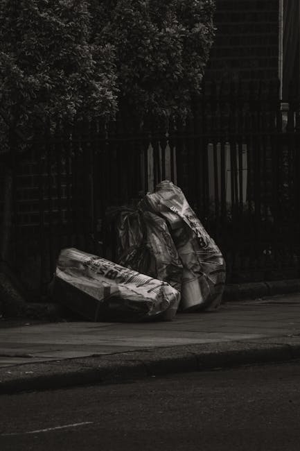 Three black plastic garbage bags filled with waste are situated on the pavement in front of a dark wooden fence and dense bushes with leafy foliage. The bags appear to be full and are arranged with two leaning against the fence and one lying flat on the sidewalk. The surface of the bags shows visible creases and some text, indicating they are used for waste disposal. The background includes the shadowed fence and greenery, suggesting an outdoor setting near residential or commercial premises where waste collection or private rubbish removal might occur. The overall scene conveys a typical collection of refuse awaiting pickup, associated with waste disposal services such as those provided by Waste Disposal Chiswick, highlighting the process of on-site rubbish clearing.