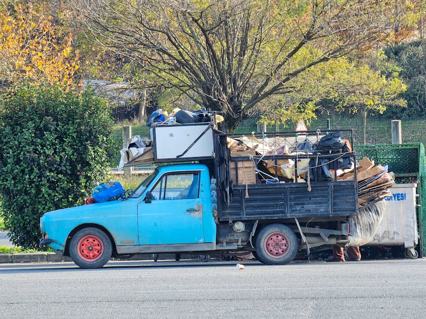 A small, light blue utility truck parked on the side of a street, heavily loaded with assorted waste materials. The cargo area is filled with a mixture of flattened cardboard boxes, black plastic bags, and various debris, secured with ropes and straps. The vehicle's tires are red, and the truck appears to be aged, with visible signs of wear on the paintwork. In the background, there is a large tree with green and yellow leaves, indicating an outdoor environment during autumn, and a metal fence surrounding a park or garden area. The scene suggests an occasion of on-site rubbish clearance or private waste collection, potentially managed by waste disposal services such as Waste Disposal Chiswick, to handle large or non-standard waste loads outside regular council collection services. The natural daylight and clear weather conditions highlight the industrial and functional setting typical for rubbish removal activities.