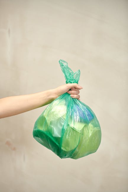 A person's hand is holding a green and blue plastic rubbish bag by its twisted, tightly knotted top against a plain, neutral beige background. The bag appears to contain lightweight waste materials, possibly soft plastics or household refuse, and is slightly bulging, indicating it is partially filled. The person's arm and hand are visible, with the arm extending from the left side of the image, and the skin tone is light. There are no other objects or environmental details visible in the image. This scene relates to waste disposal practices, suggesting an act of rubbish collection or disposal, which is relevant to professional waste management services such as those provided by Waste Disposal Chiswick, operating within the scope of private or alternative rubbish removal options. The lighting is neutral and even, emphasizing the colours and textures of the plastic bag without creating shadows or reflections.