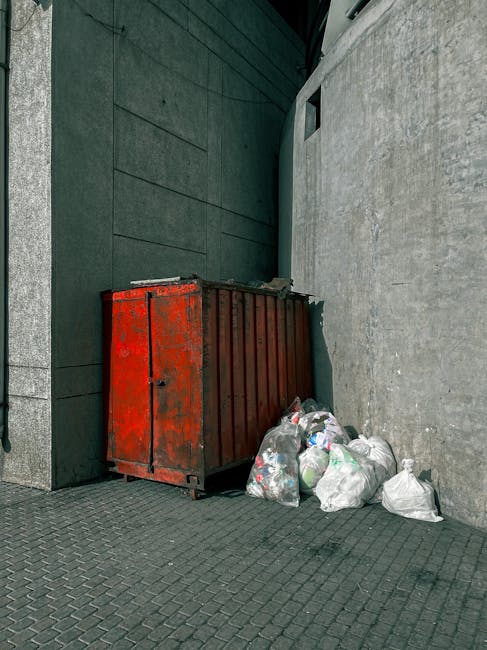A weathered red metal skip with visible rusting and peeling paint positioned against a dark green wall and a light grey concrete wall in an urban exterior setting. The skip is situated on a paved surface and is partially overflowing with several white plastic rubbish bags filled with mixed waste. Additional bags are piled on the ground beside the skip, leaning against it, and appear loosely tied. The environment appears to be a narrow alleyway or backstreet area, with the scene illuminated by natural daylight. This setting illustrates a typical private waste collection scenario, where external waste receptacles are used for rubbish disposal, aligning with methods of alternative waste handling and rubbish removal services such as those provided by Waste Disposal Chiswick.