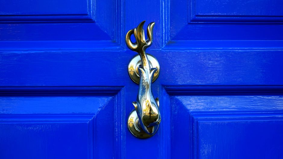 A close-up view of a bright blue wooden door with a glossy finish, featuring four rectangular recessed panels. Mounted on the door is a polished brass door handle plate with a traditional round rosette, to which a decorative brass door knocker shaped like a dog's head is attached. The door knocker has a smooth, shiny surface that reflects light, and a curved tongue that serves as the knocking element. The scene is evenly lit, with reflections highlighting the metallic surfaces, and the blue paint surrounding the fixtures shows distinct wood grain and panel edges. This image pertains to private property access, relevant to waste disposal or rubbish removal services, as part of alternative waste handling solutions where secure entry points are necessary for on-site clearance by professional waste disposal companies like Waste Disposal Chiswick.