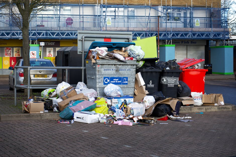 A large collection of mixed waste is overflowing from a grey recycling bin labelled for paper and card, positioned on a paved area in front of a commercial building. The pile includes crumpled cardboard boxes, plastic bags, paper packaging, and other discarded items, some spilling onto the nearby curb and sidewalk. To the right of the recycling bin, there are black and red waste containers, also filled with rubbish, with additional bags and loose waste scattered around their bases. A small silver car is parked behind the waste despite a metal railing separating vehicles from the rubbish area. The background features a multi-storey building with blue scaffolding and a storefront with signage, indicating a commercial or retail environment. The scene is captured during daylight hours, with natural light illuminating the cluttered waste and surrounding urban setting. This image illustrates the type of visual scenario that waste disposal services like those provided by Waste Disposal Chiswick handle, particularly when dealing with unmanaged or improperly contained rubbish in public spaces.